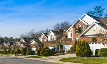 Row of brick suburban houses with manicured bushes and driveways, some with parked cars, on a sunny day under a blue sky. Trees and clear sidewalks line the residential street.