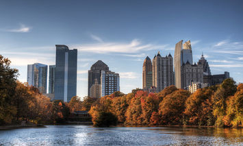 Skyscrapers standing tall against a blue sky, framed by autumn-colored trees with a calm lake in the foreground.