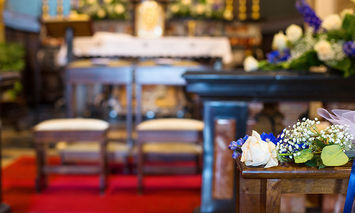 A small floral bouquet rests on a wooden pew against a background of an elaborately decorated church interior with an altar, floral arrangements, and candles, all on a red carpet.