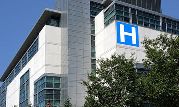 A multi-story hospital building with large windows displays a blue sign with a white 