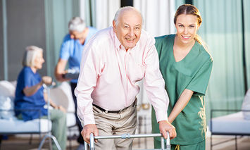 An elderly man using a walker is assisted by a smiling nurse in green scrubs, while other elderly individuals are engaged in activities in an outdoor, sunlit care facility setting.