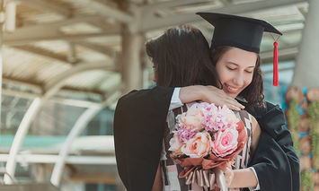 A graduate wearing a cap and gown hugs someone while holding a bouquet of flowers in an outdoor setting with arched architecture in the background.